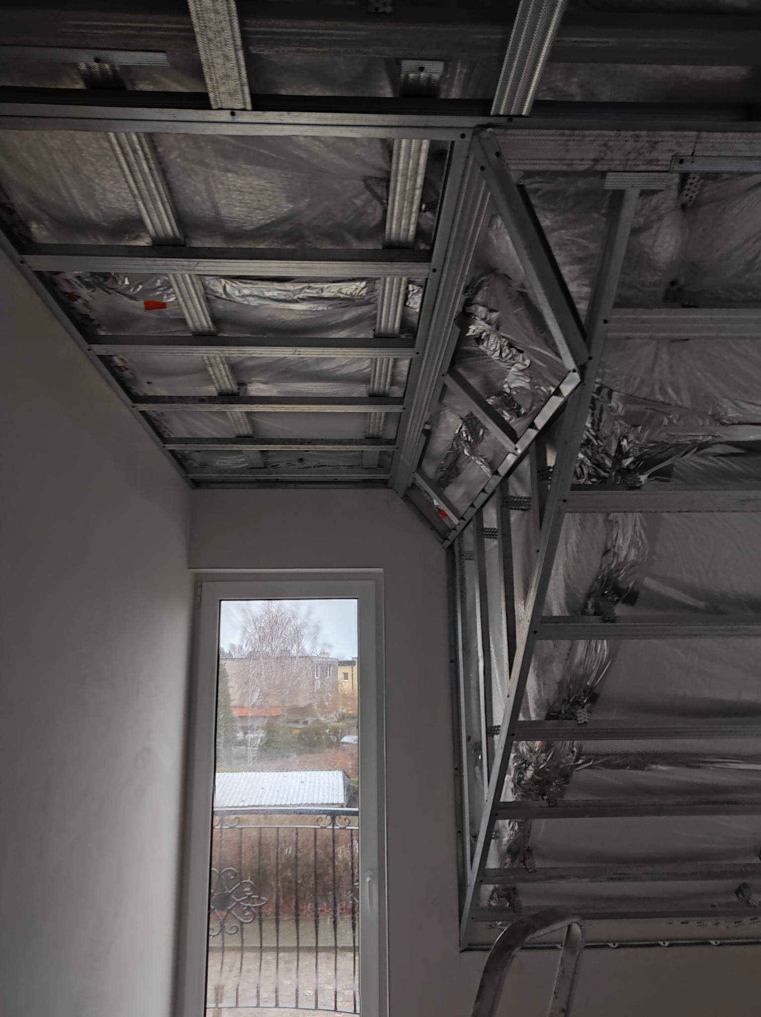 Stelaż metalowy pod płyty karton-gips z izolacją folią aluminiową, widok na okno z widokiem na ogród i budynki w tle.