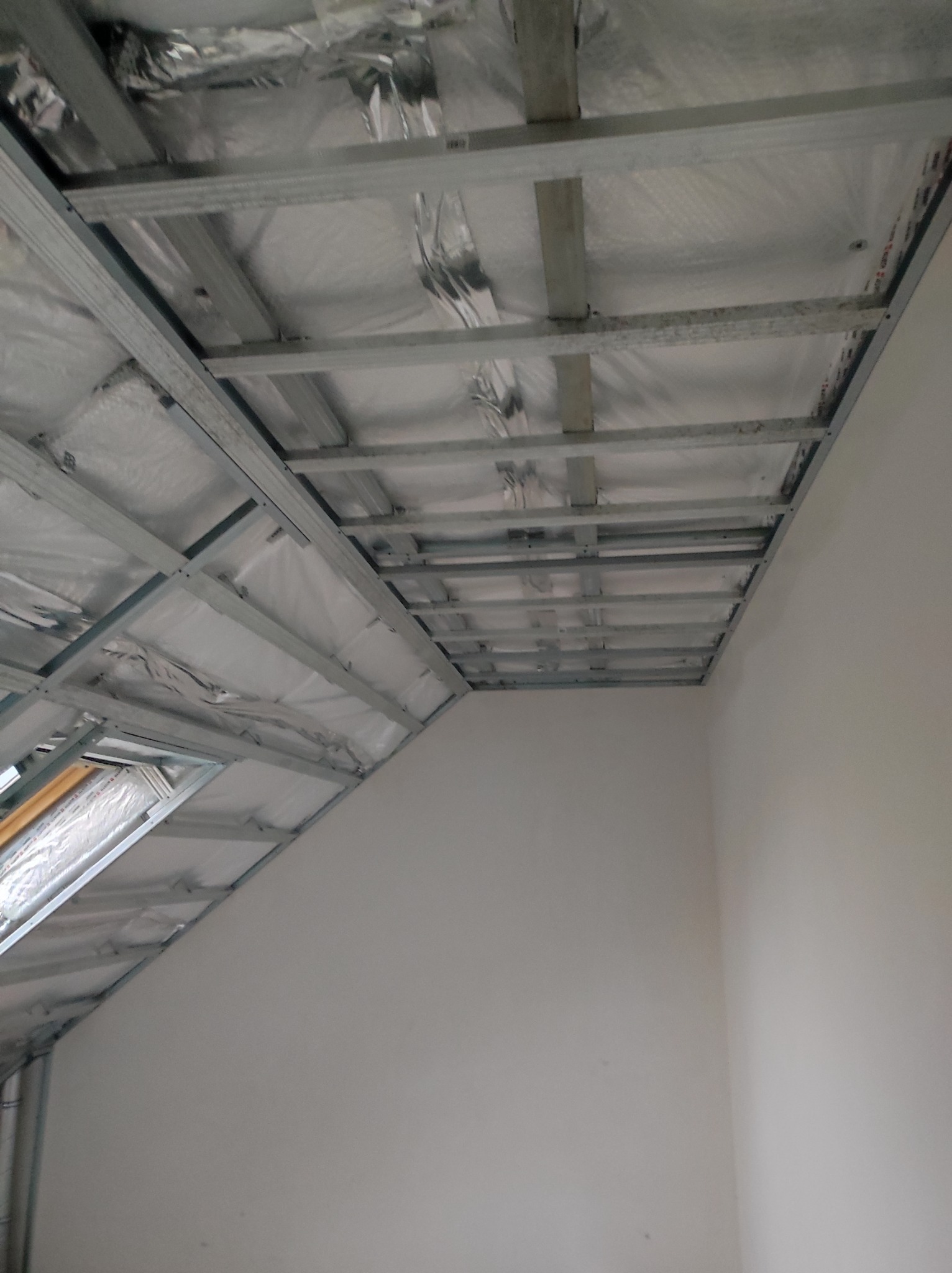 Szkielet metalowy pod konstrukcję sufitu podwieszanego na poddaszu, widoczna izolacja z folii aluminiowej, okno dachowe w lewym dolnym rogu.
