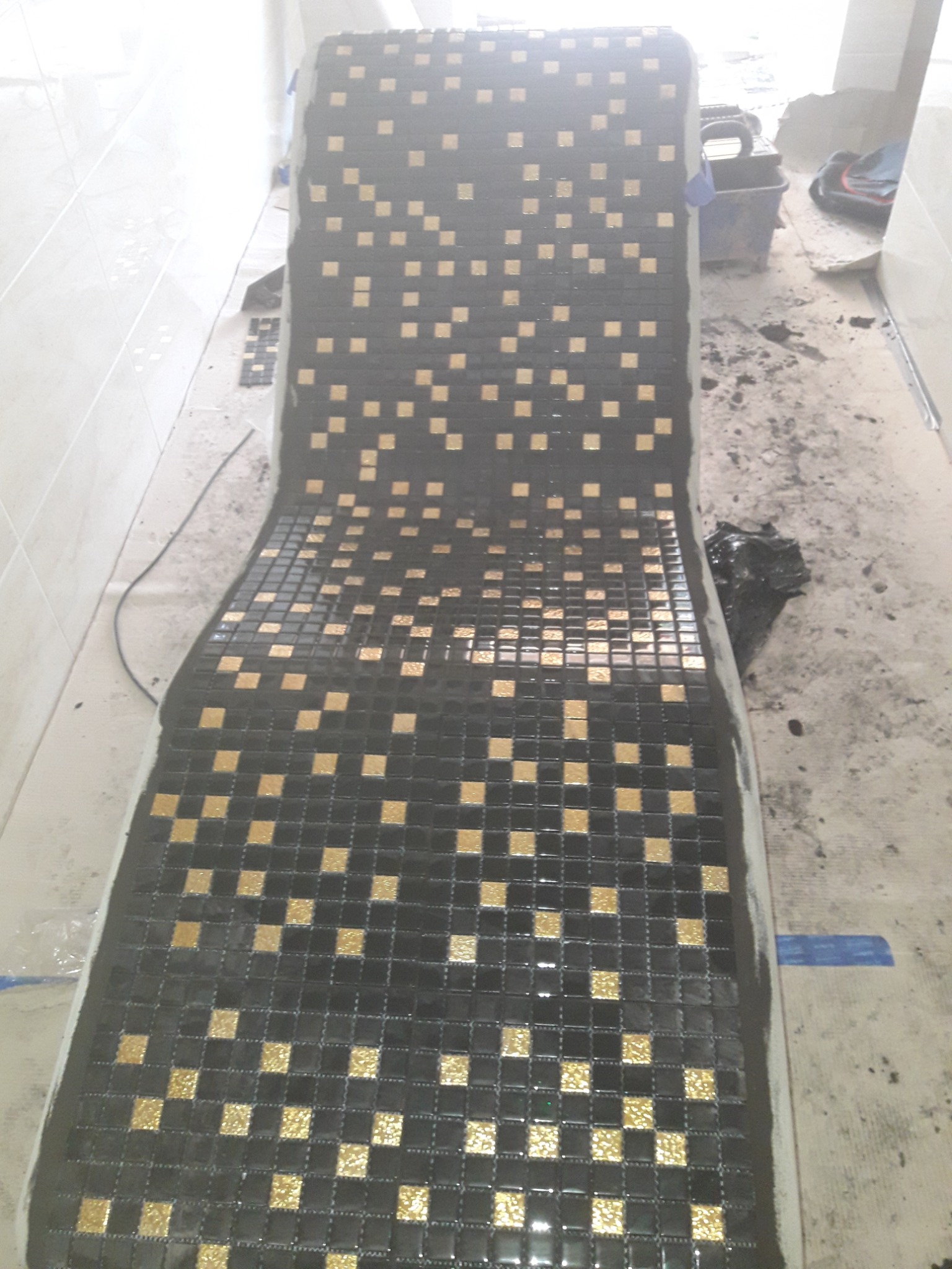 Układanie mozaiki w łazience, czarne i złote kwadratowe płytki tworzące wzór na zakrzywionej powierzchni, widoczne narzędzia i resztki zaprawy na podłodze.