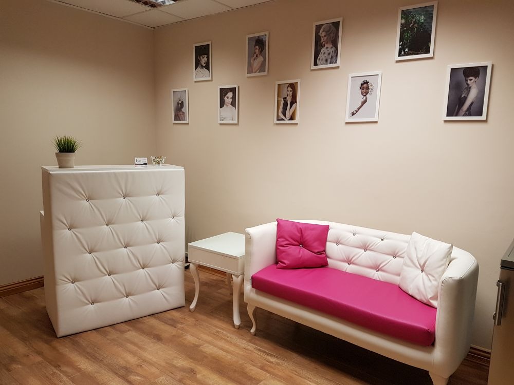 Jasne wnętrze salonu: biała, pikowana lada recepcyjna z zieloną rośliną, kanapa z różowym siedziskiem i poduszkami, zdjęcia portretowe na ścianie.