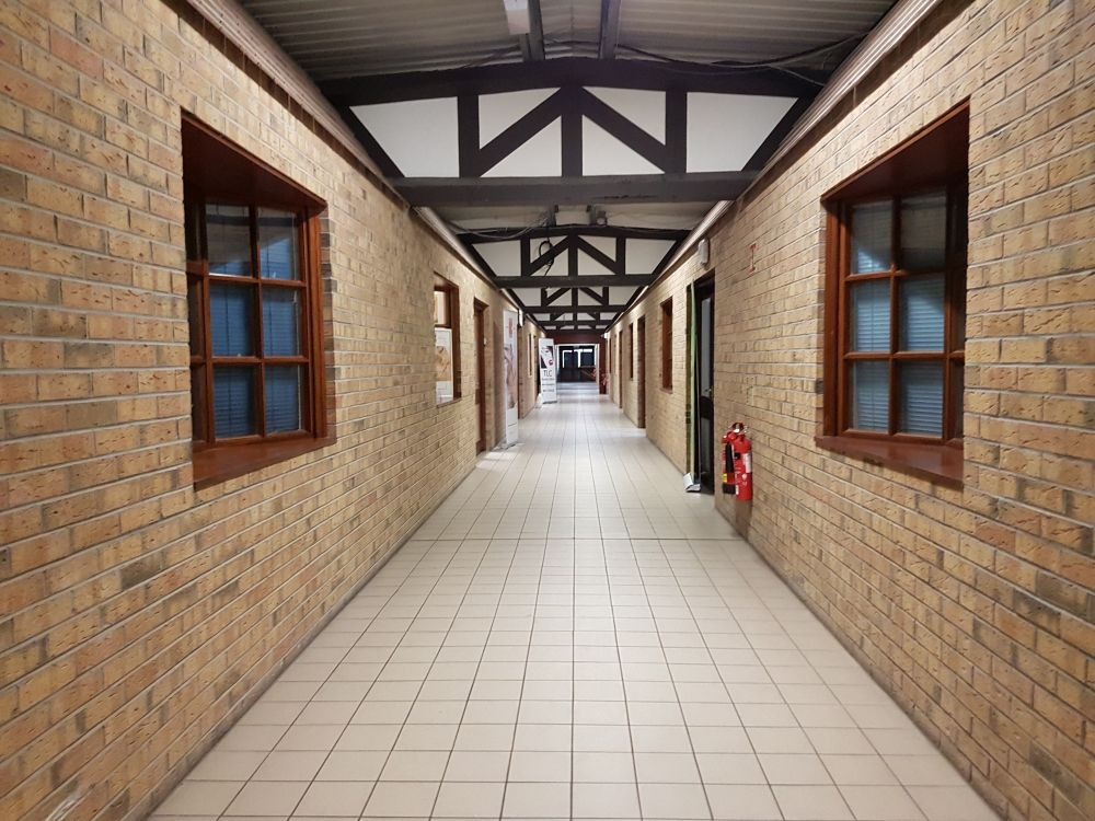 Długi korytarz z ceglanymi ścianami, drewnianymi oknami i białą kafelkową podłogą, perspektywa zbieżna ku końcowi korytarza, widoczna gaśnica na ścianie.