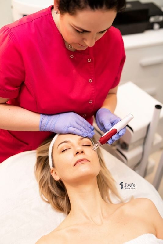 Kosmetolog w różowej bluzie, w niebieskich rękawiczkach wykonuje zabieg mezoterapii igłowej na twarzy klientki leżącej na białym łóżku kosmetycznym, widoczne logo salonu w prawym dolnym rogu.