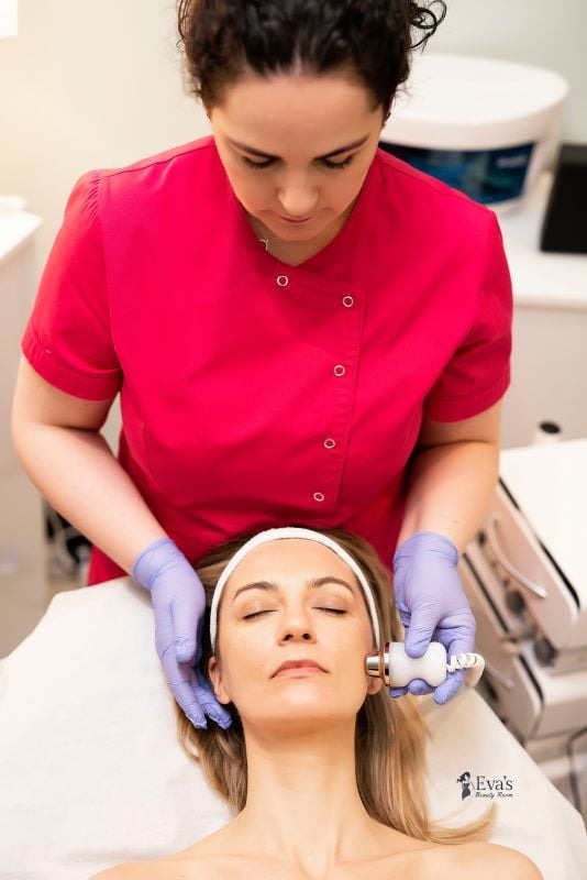 Kobieta w różowym uniformie i niebieskich rękawiczkach wykonuje zabieg na twarzy klientki leżącej na białym łóżku kosmetycznym, używając urządzenia z metalową końcówką.