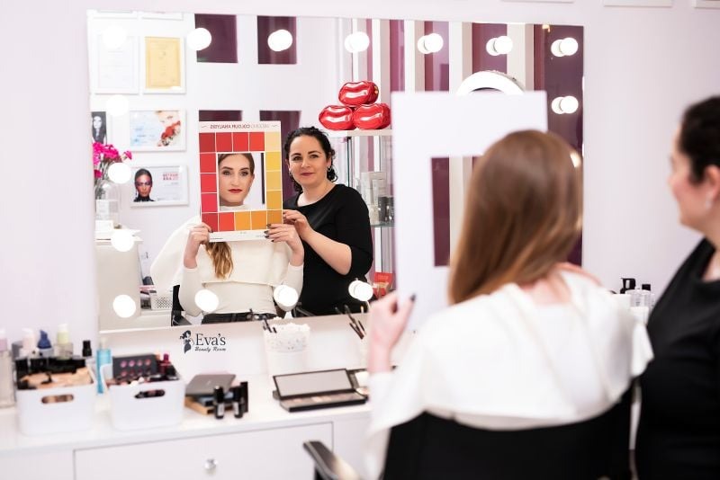 Klientka prezentuje twarz w ramce z paletą kolorów do makijażu, konsultantka pomaga w doborze odcieni, w tle lustro z oświetleniem i kosmetyki.