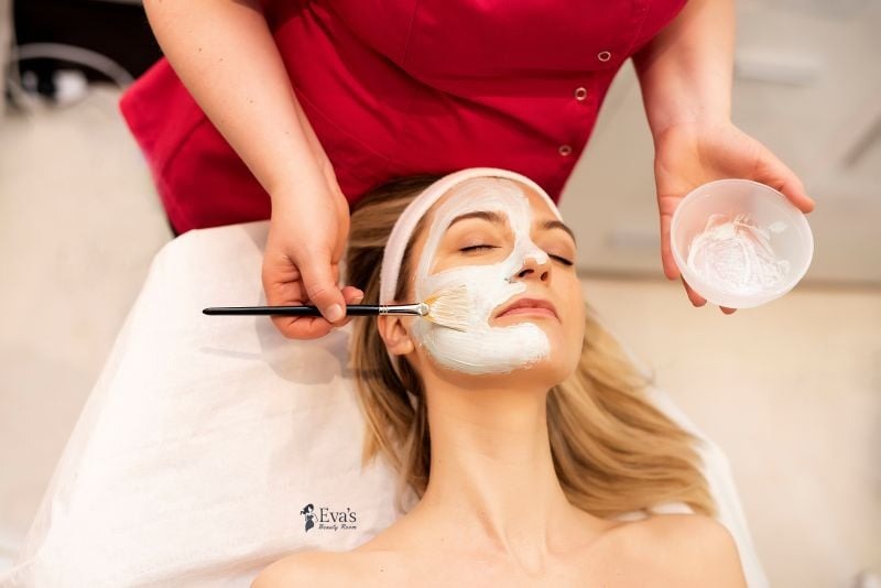Nakładanie białej maski na twarz klientki pędzlem w salonie kosmetycznym, widoczna miseczka z maską.