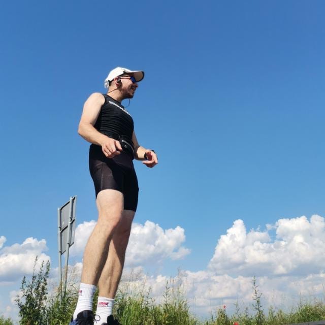 Uśmiechnięty mężczyzna w sportowym stroju, z czapką i słuchawkami, stoi na tle jasnego nieba z chmurami, prawdopodobnie przed biegiem lub po nim.