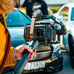 Kamera na stabilizatorze w rękach operatora, w tle dwa samochody i osoby w czarnych ubraniach, ujęcie od strony operatora.