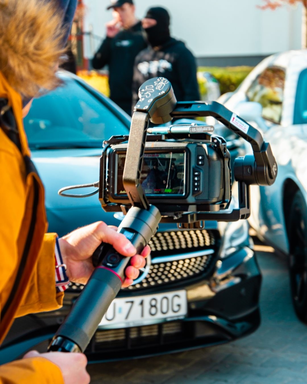 Kamera na stabilizatorze w rękach operatora, w tle dwa samochody i osoby w czarnych ubraniach, ujęcie od strony operatora.