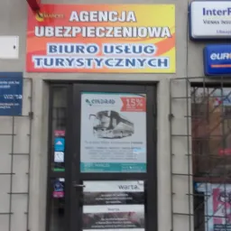 Fasada budynku agencji ubezpieczeniowej i biura usług turystycznych z widocznymi szyldami reklamowymi firm ubezpieczeniowych InterRisk i Warta oraz przewoźnika autokarowego EuroLines i Sindbad.
