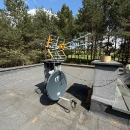 Antena telewizyjna i satelitarna Invertio zamontowane na dachu budynku, z lasem w tle i błękitnym niebem. Widoczne kable połączeniowe.