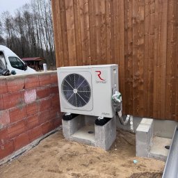 ELTROCOM SP. Z O.O. - Biała pompa ciepła Rotenso Aquami zamontowana na betonowych podstawach przy drewnianej ścianie budynku. Widoczne rury i elementy instalacji.