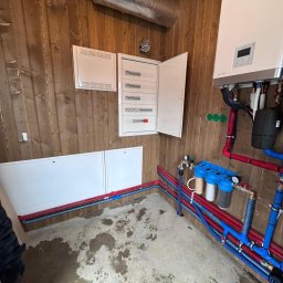 ELTROCOM SP. Z O.O. - Rozdzielnia elektryczna z otwartymi drzwiami, biały bojler z podłączeniami hydraulicznymi i filtry wody na tle boazerii. Widoczne rury w kolorze czerwonym i niebieskim.