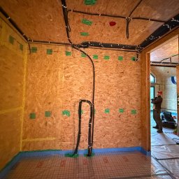 ELTROCOM SP. Z O.O. - Wnętrze domu w trakcie prac: widoczne kable elektryczne prowadzone po ścianach z płyt OSB, instalacja ogrzewania podłogowego. W tle majster.