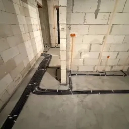 Szare rury instalacji elektrycznej ułożone na podłodze w surowym wnętrzu z białymi ścianami z betonu komórkowego i pomarańczowymi puszkami elektrycznymi.