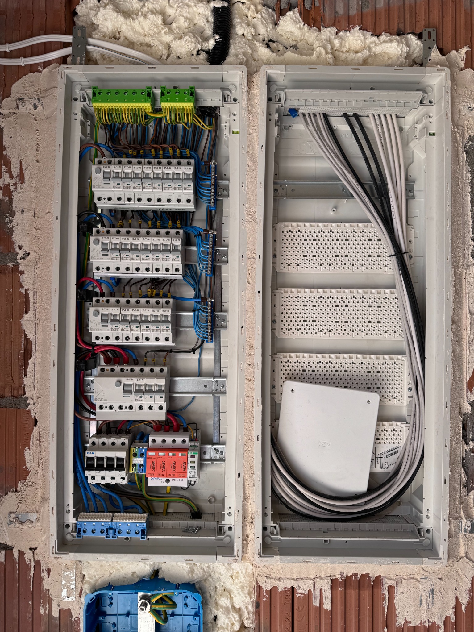 Rozdzielnica elektryczna w trakcie montażu w surowej ścianie z cegły, widoczne kable, bezpieczniki, listwy zaciskowe i piana montażowa.