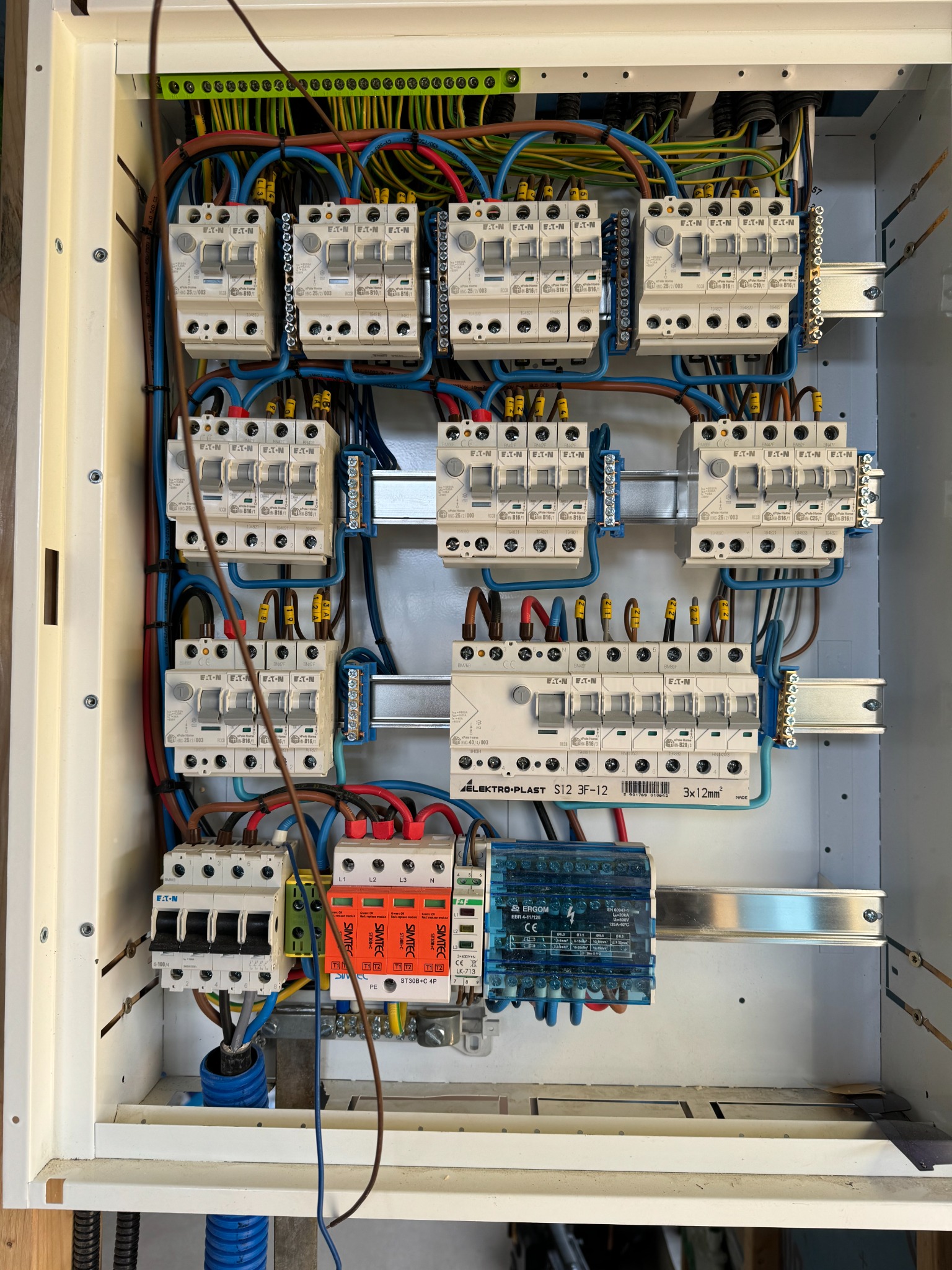 Otwarta skrzynka rozdzielcza z zamontowanymi bezpiecznikami EATON, zabezpieczeniami SIMTEC i modułem ERGOM, widoczne okablowanie, szyny montażowe i elementy mocujące.