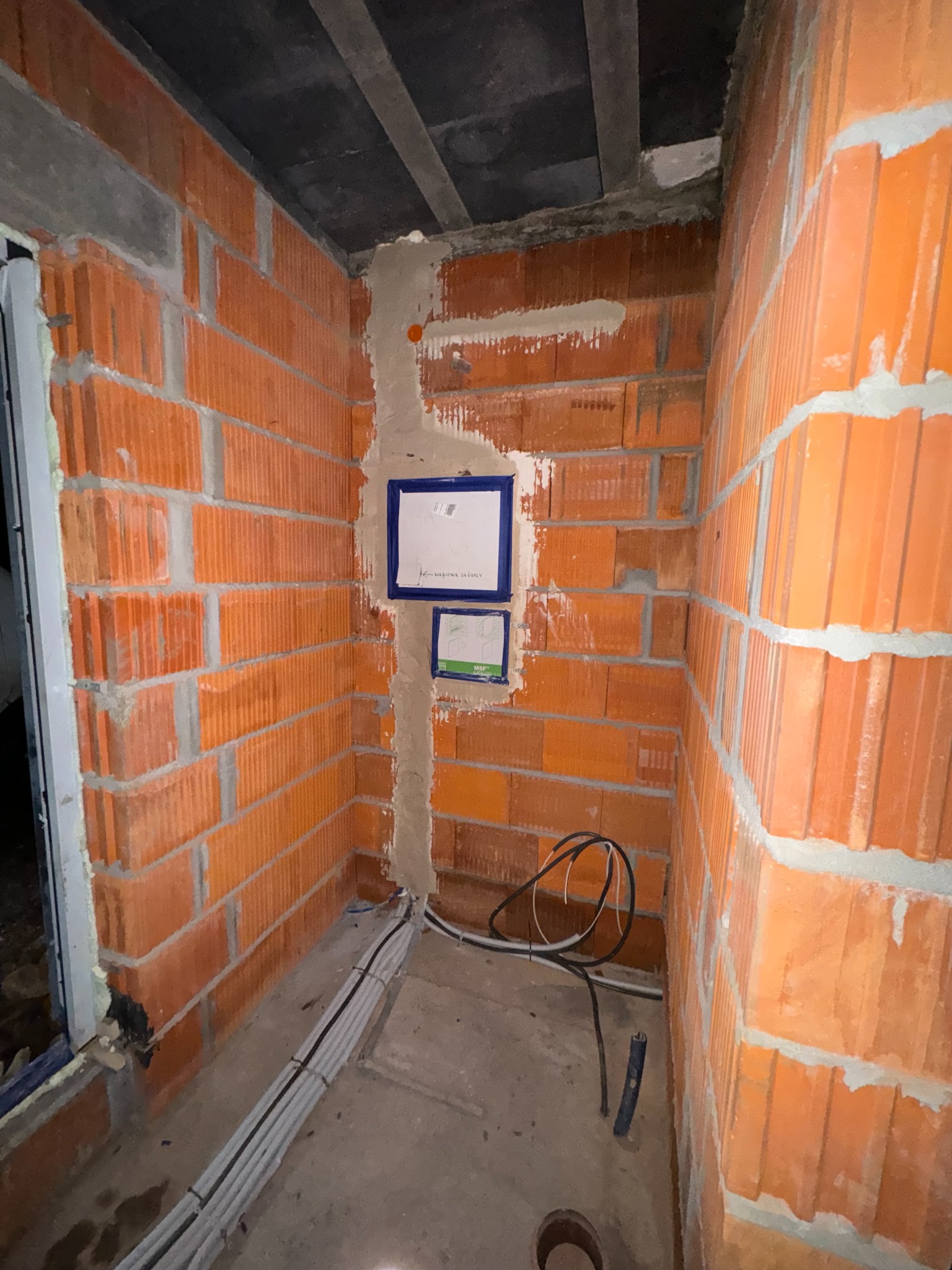 Surowy mur z cegły dziurawki, częściowo otynkowany, z widocznymi przewodami elektrycznymi i puszkami podtynkowymi, przygotowany do dalszych prac wykończeniowych.