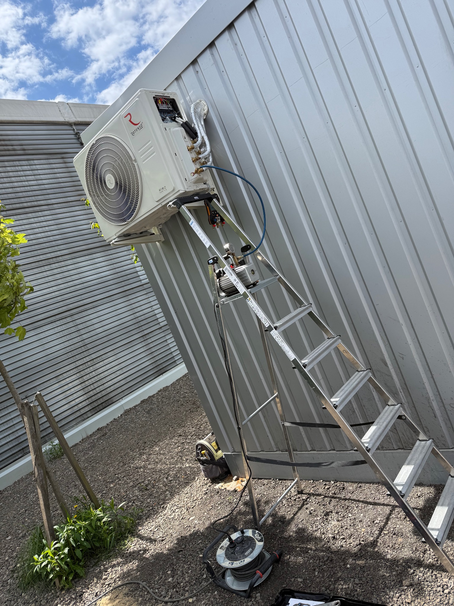 Biała jednostka zewnętrzna klimatyzacji Rotenso zamontowana na metalowej ścianie, dostępna po drabinie. Widoczne narzędzia i przewody sugerują trwający montaż.