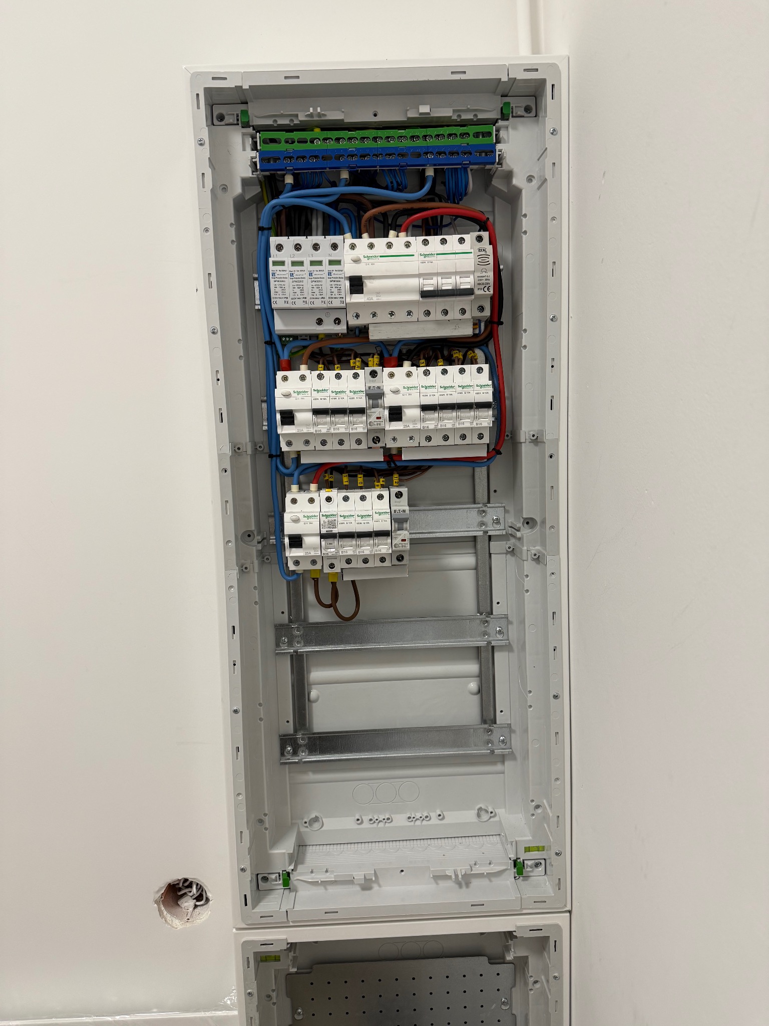 Rozdzielnica elektryczna w obudowie, widoczne bezpieczniki, przewody w kolorach: niebieskim, czerwonym, brązowym i żółto-zielonym. Instalacja w trakcie montażu. Otwór w ścianie obok.