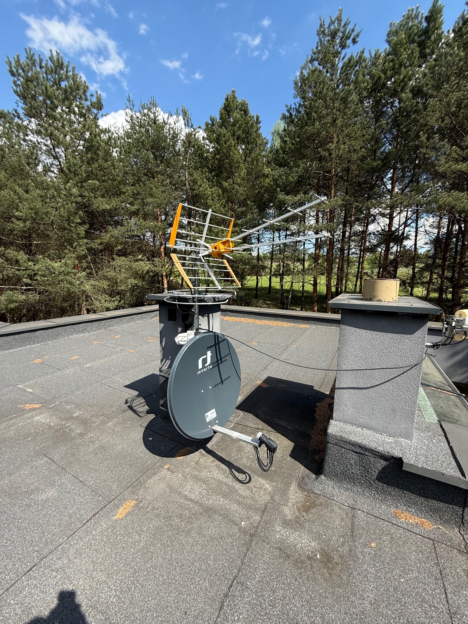Antena telewizyjna i satelitarna Invertio zamontowane na dachu budynku, z lasem w tle i błękitnym niebem. Widoczne kable połączeniowe.