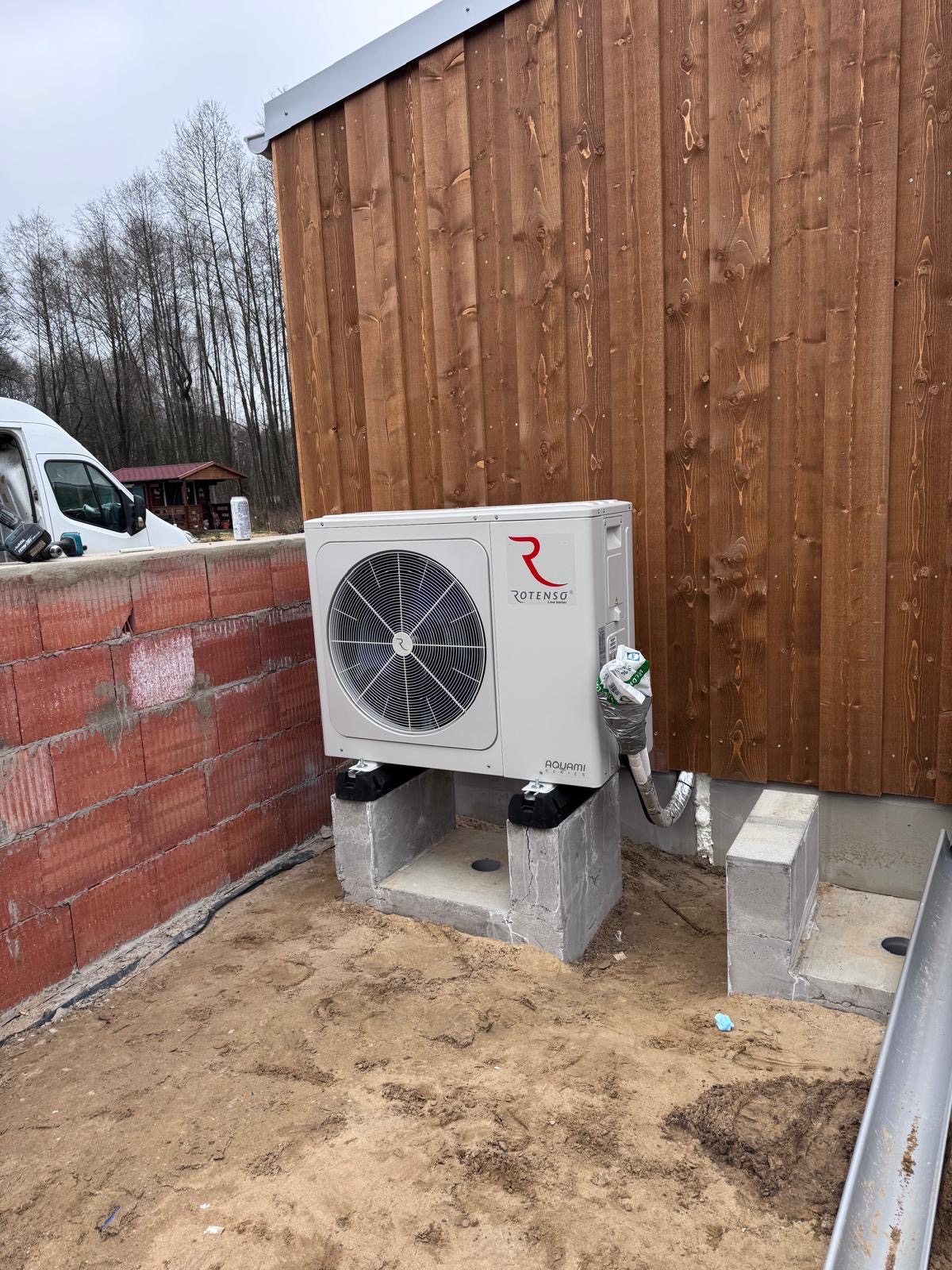 Biała pompa ciepła Rotenso Aquami zamontowana na betonowych podstawach przy drewnianej ścianie budynku. Widoczne rury i elementy instalacji.