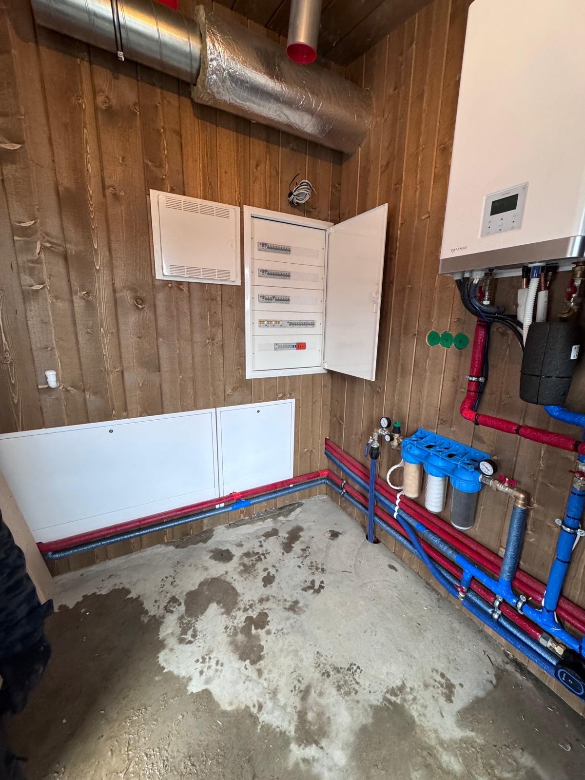 Rozdzielnia elektryczna z otwartymi drzwiami, biały bojler z podłączeniami hydraulicznymi i filtry wody na tle boazerii. Widoczne rury w kolorze czerwonym i niebieskim.