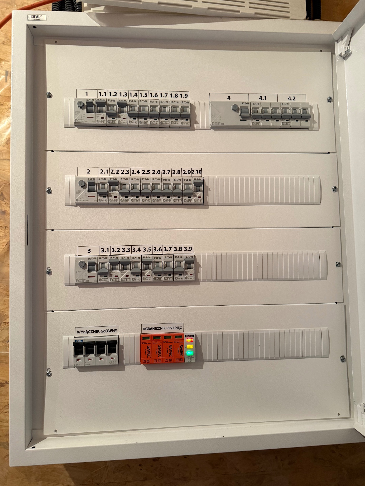 Otwarta, biała skrzynka rozdzielcza z zainstalowanymi bezpiecznikami EATON i ogranicznikiem przepięć Schelinger. Widoczne oznaczenia numeracji obwodów.
