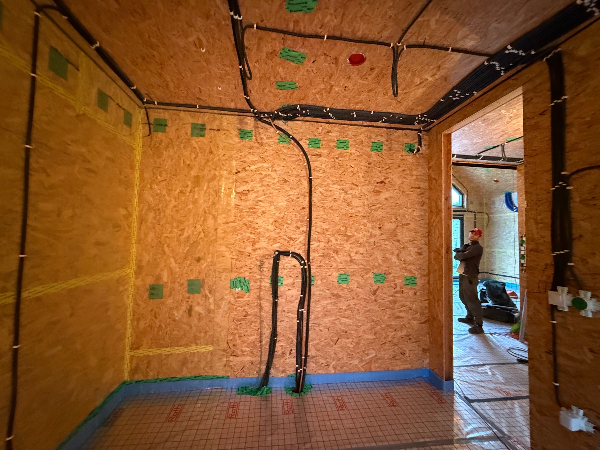Wnętrze domu w trakcie prac: widoczne kable elektryczne prowadzone po ścianach z płyt OSB, instalacja ogrzewania podłogowego. W tle majster.