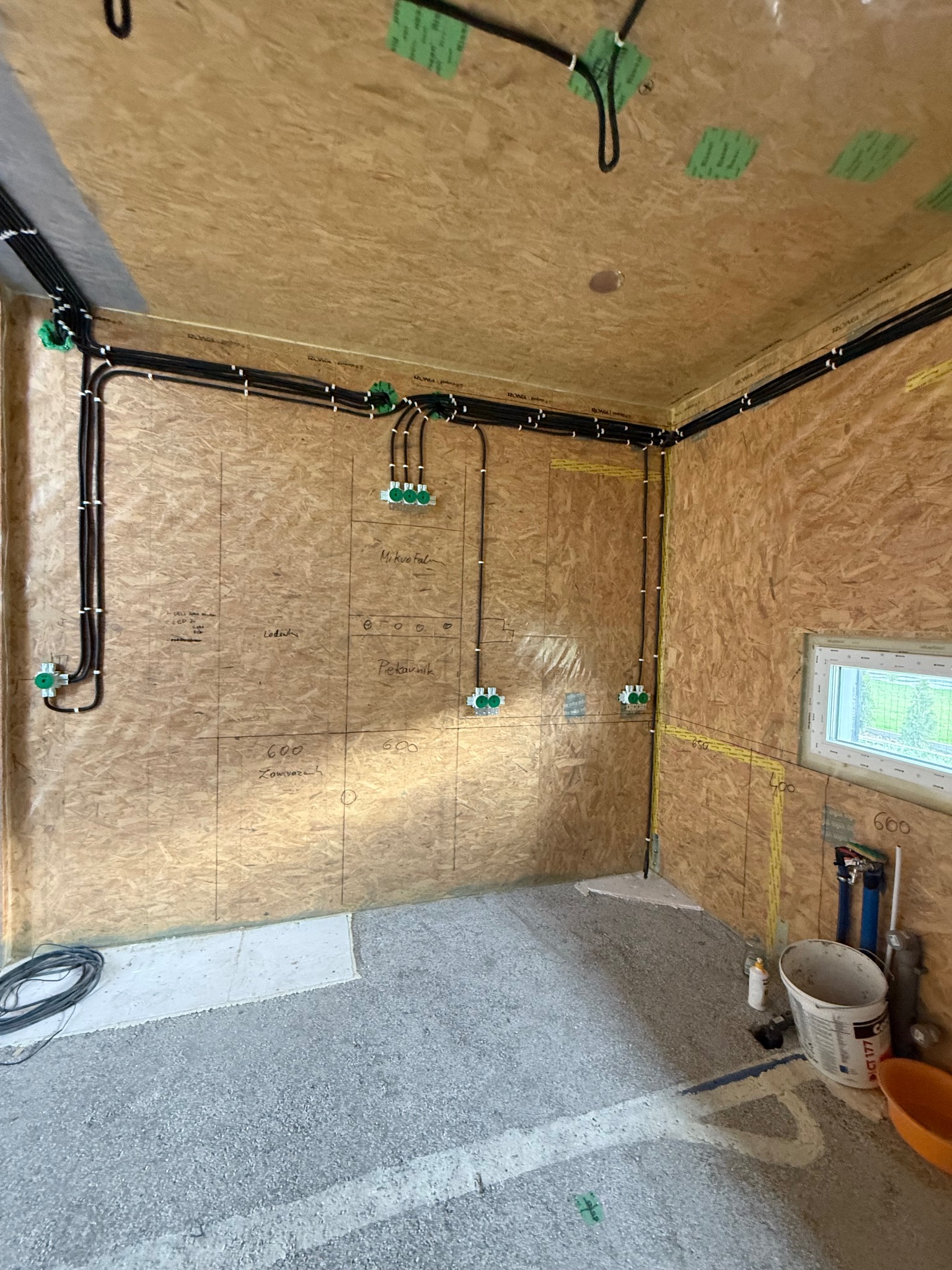Wnętrze w trakcie instalacji elektrycznej: kable w peszlach, puszki podtynkowe, ściany wyłożone płytami OSB z naniesionymi odręcznymi oznaczeniami, surowa wylewka.
