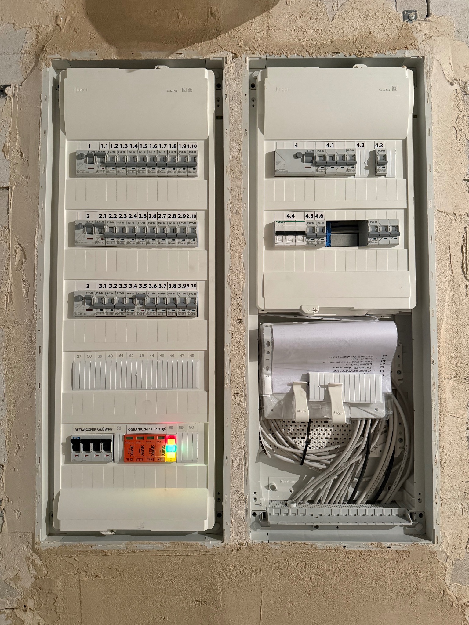 Rozdzielnica elektryczna w trakcie montażu, widoczne kable, zabezpieczenia i schemat. Częściowo odkryta ściana w tle. Tablica z bezpiecznikami.
