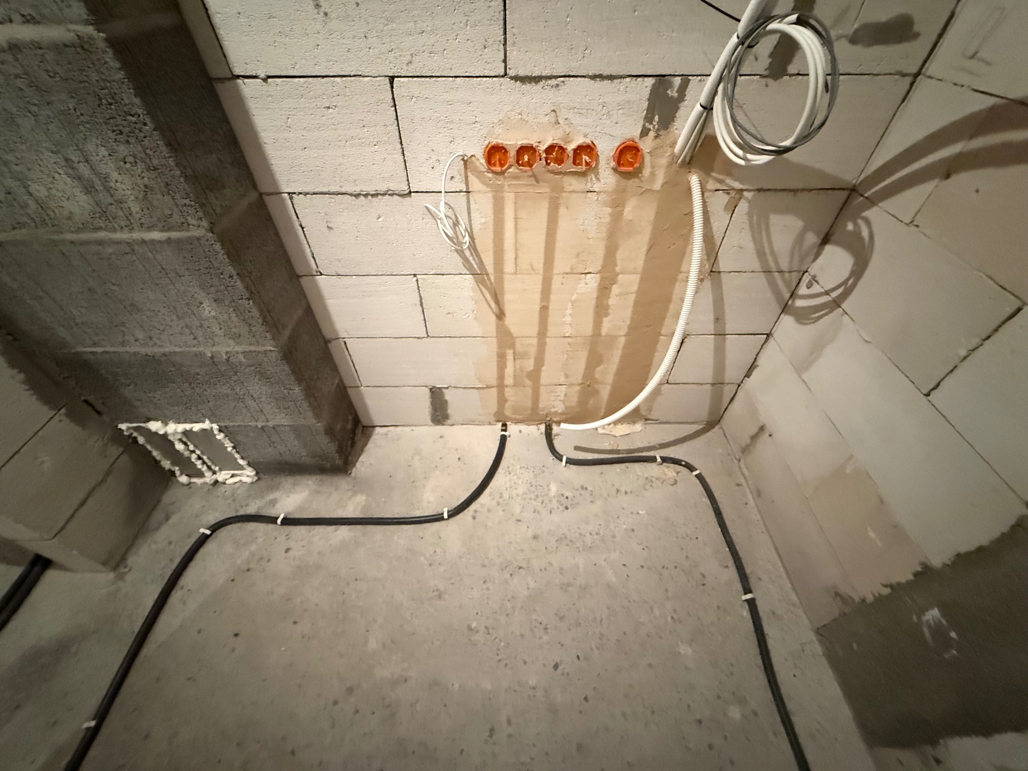 Surowy stan: Instalacja elektryczna w ścianie z bloczków, widoczne kable i puszki. Elementy mocowane do ściany za pomocą zaprawy. Kable ułożone wzdłuż podłogi.