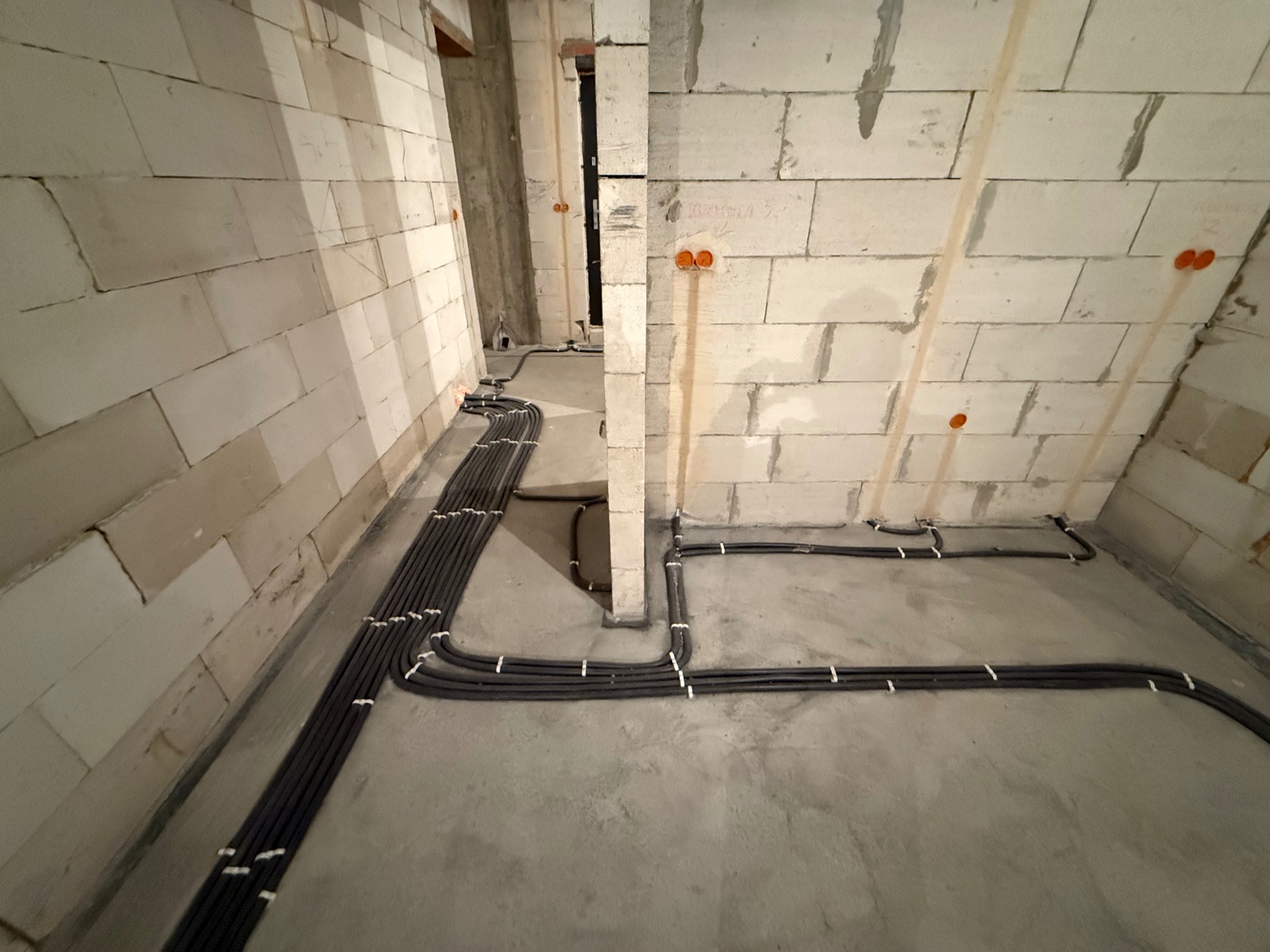 Szare rury instalacji elektrycznej ułożone na podłodze w surowym wnętrzu z białymi ścianami z betonu komórkowego i pomarańczowymi puszkami elektrycznymi.