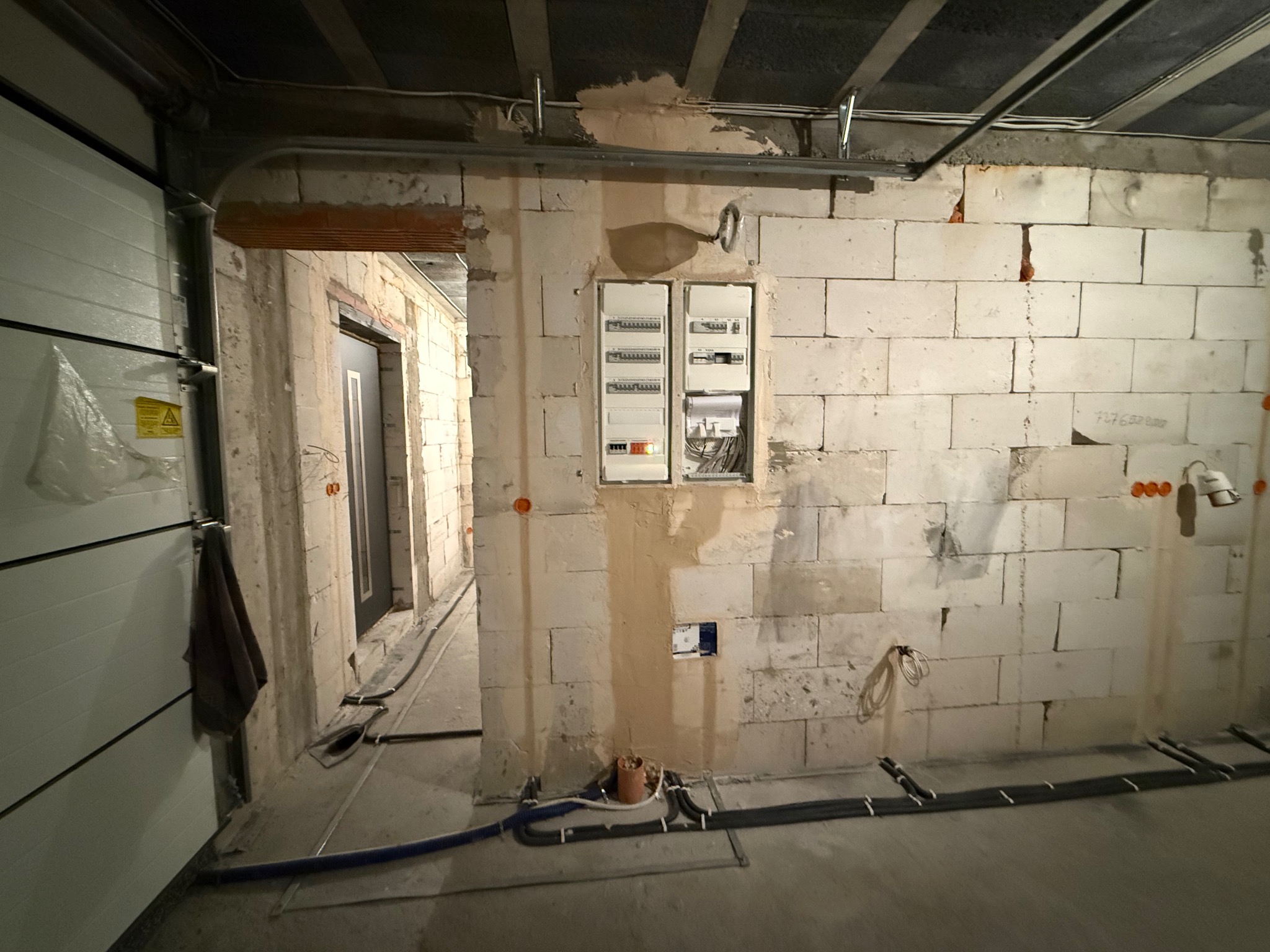 Wnętrze w trakcie prac: ściana z bloczków, rozdzielnica elektryczna wnękowa, rury instalacyjne na podłodze, widoczne okablowanie. Surowy stan, etap prac instalacyjnych.