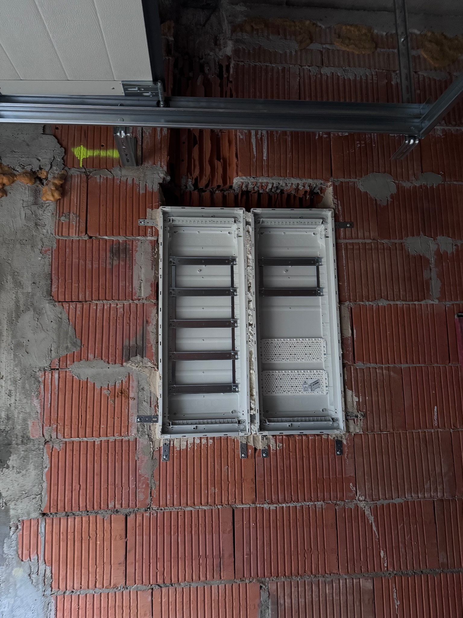 Wnęka w surowej, ceglanej ścianie z zamontowaną, pustą rozdzielnicą elektryczną. Widoczne elementy konstrukcyjne sufitu. Żółta strzałka namalowana na ścianie.