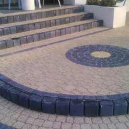 Schody i okrąg na posesji wyłożone kostką brukową w odcieniach szarości i beżu, z czarną obwódką, widoczne z bliska w słoneczny dzień.