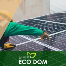 Pracownik w kasku i rękawicach montuje panel słoneczny na dachu, używając klucza. Widoczne moduły fotowoltaiczne oraz logo firmy w dolnej części kadru.