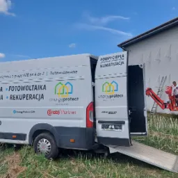 Monterska winda z pracownikiem przy białej ścianie budynku z zamontowanymi wspornikami, obok zaparkowany biały bus z reklamą firmy oferującej kompleksowe rozwiązania OZE.