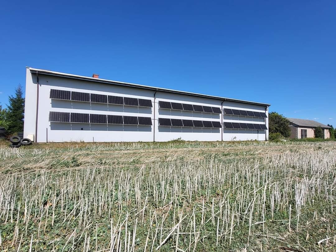 Duży budynek przemysłowy z rzędami paneli słonecznych na ścianie, widok z pola ścierniska w słoneczny dzień.