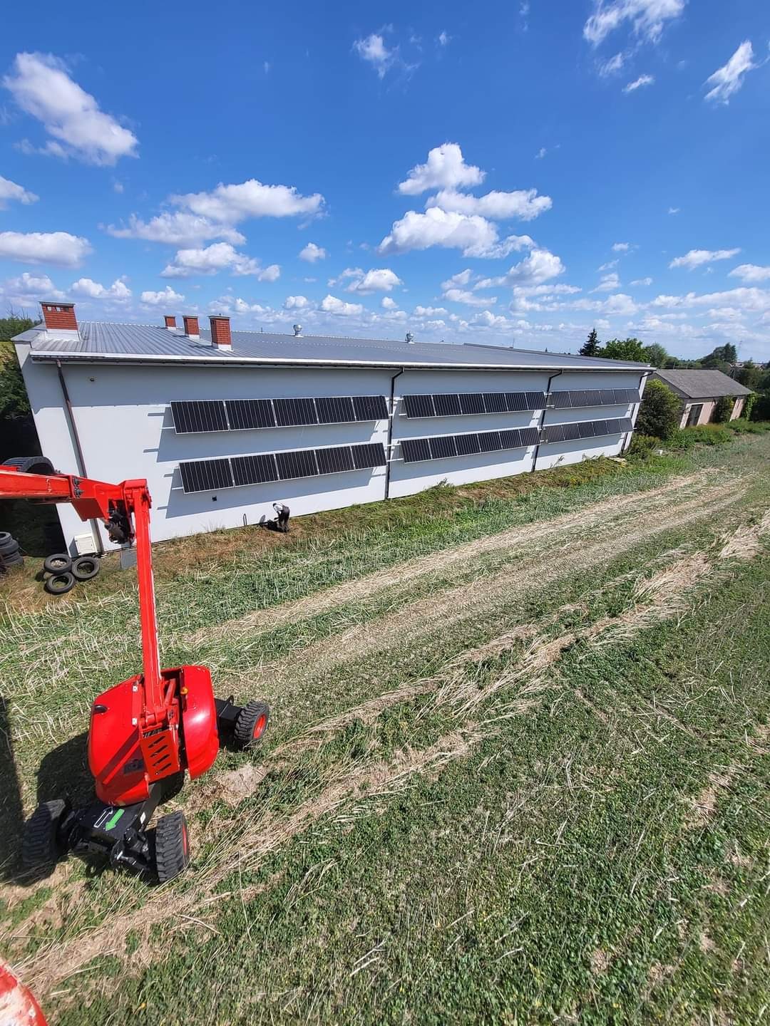 Duży budynek gospodarczy z zainstalowanymi panelami słonecznymi, widok z podnośnika koszowego na tle błękitnego nieba z chmurami.