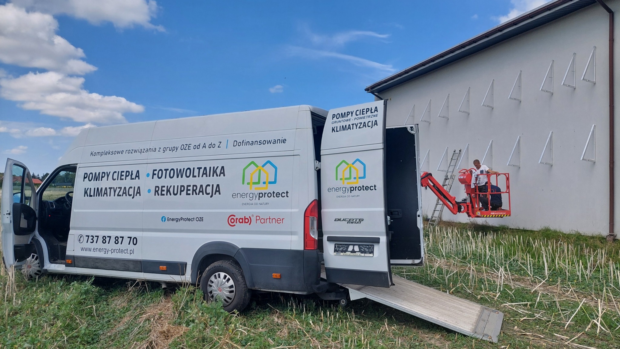 Monterska winda z pracownikiem przy białej ścianie budynku z zamontowanymi wspornikami, obok zaparkowany biały bus z reklamą firmy oferującej kompleksowe rozwiązania OZE.