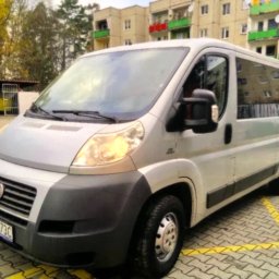 HEBAN - Srebrny bus Fiat Ducato parkuje na parkingu z żółtymi liniami, na tle bloku mieszkalnego z kolorowymi balkonami.