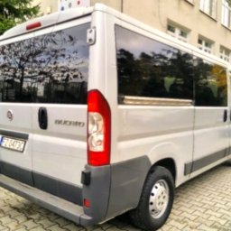 HEBAN - Srebrny bus marki Fiat Ducato, widziany z tyłu, zaparkowany na chodniku z kostki, z widocznym odbiciem drzewa w przyciemnianych szybach.