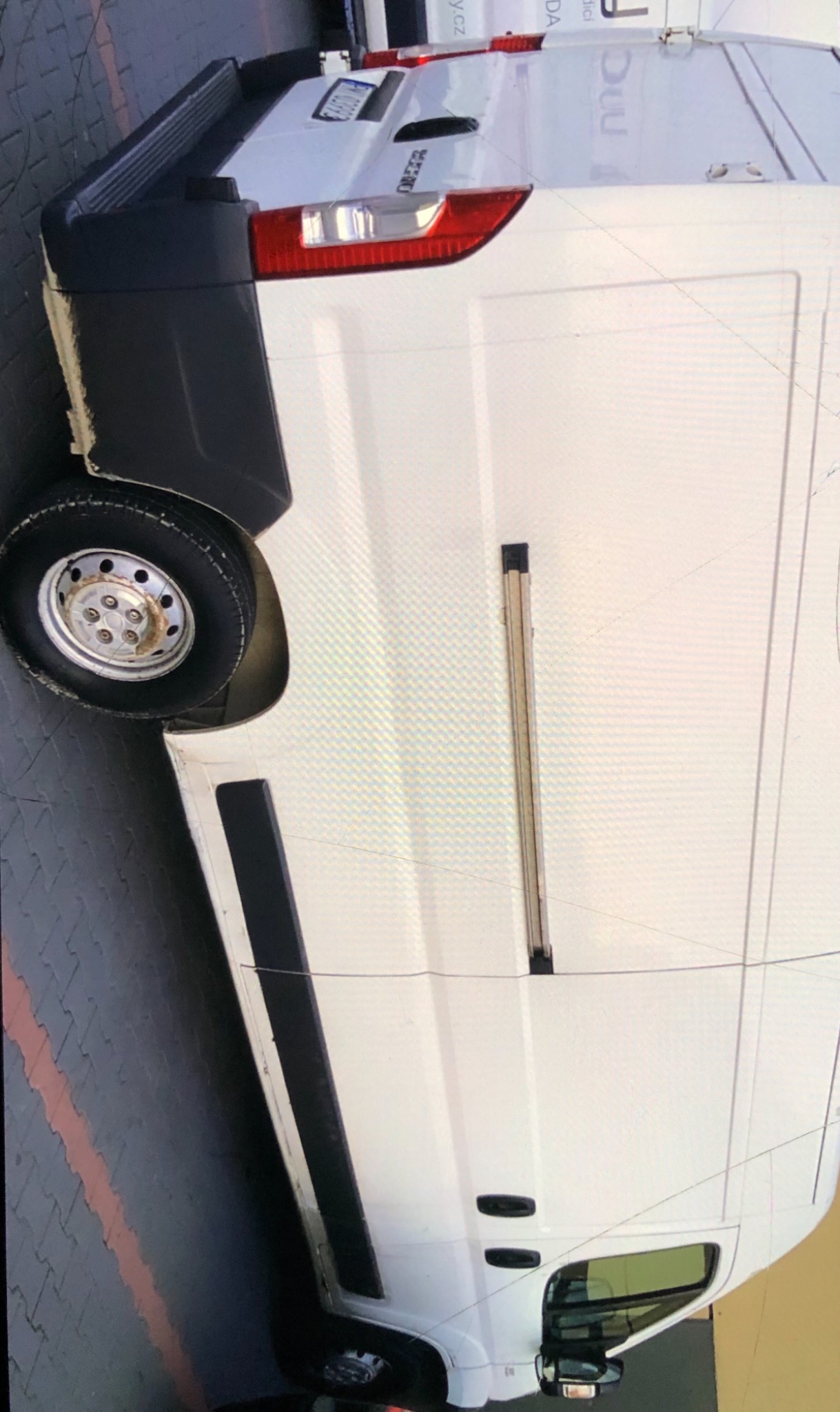 Biały bus dostawczy zaparkowany na kostce brukowej, widoczny bok pojazdu z przesuwnymi drzwiami oraz tylne światła i tablica rejestracyjna.