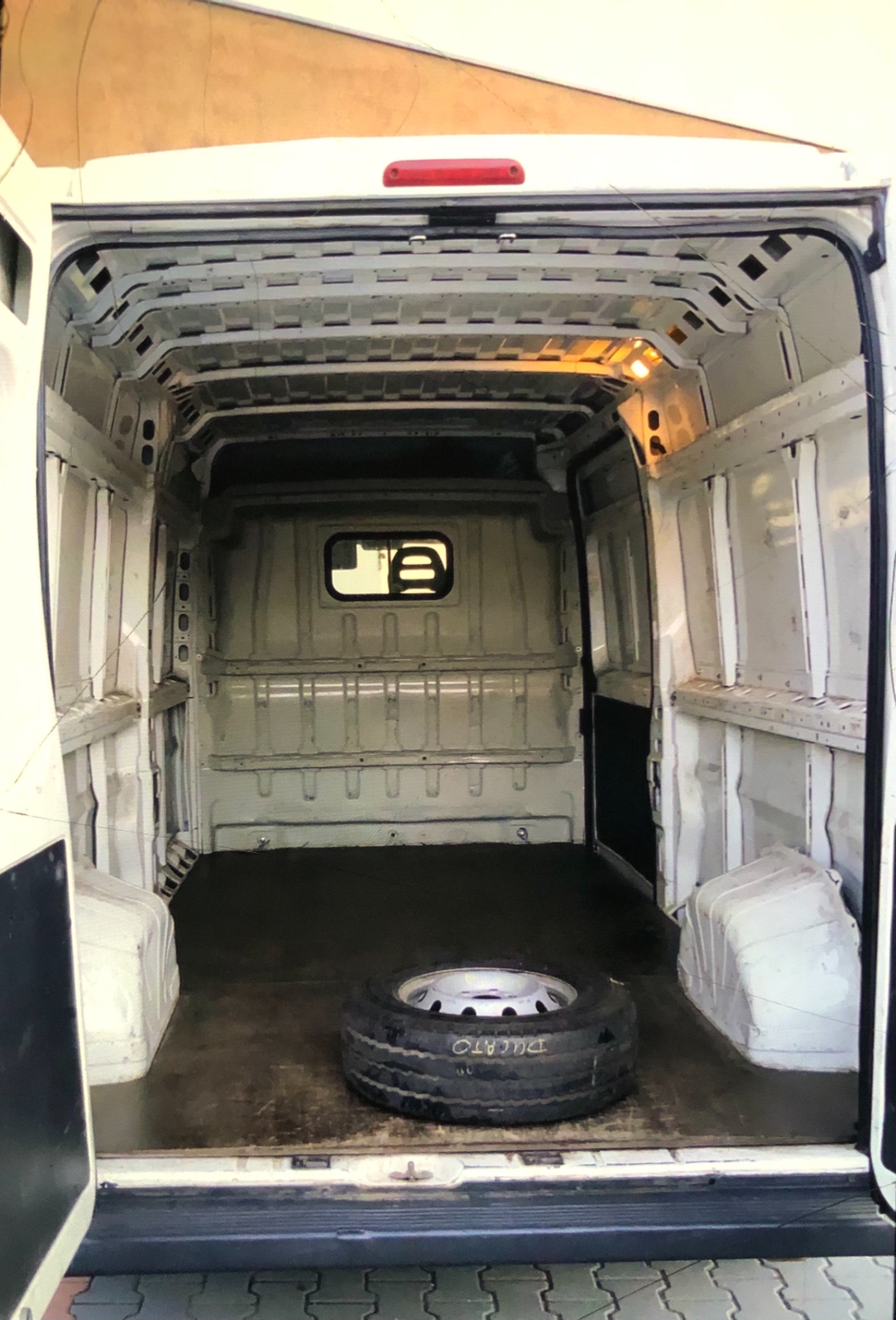 Wnętrze białego, pustego furgonetki z otwartymi tylnymi drzwiami, widoczna opona leżąca na podłodze przestrzeni ładunkowej.