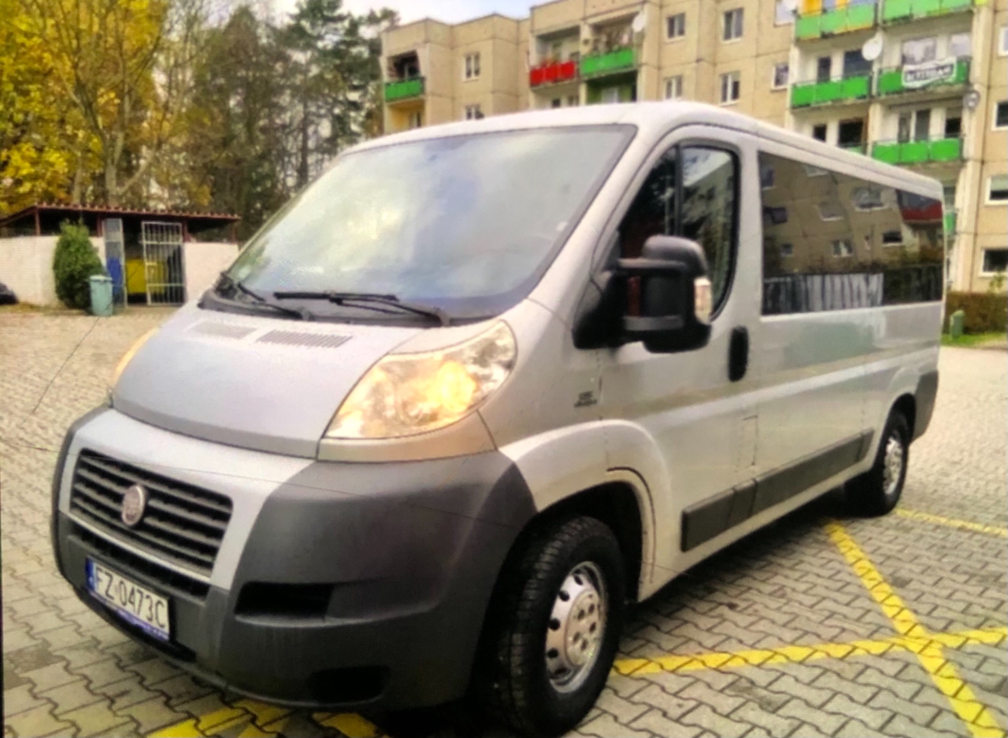 Srebrny bus Fiat Ducato parkuje na parkingu z żółtymi liniami, na tle bloku mieszkalnego z kolorowymi balkonami.
