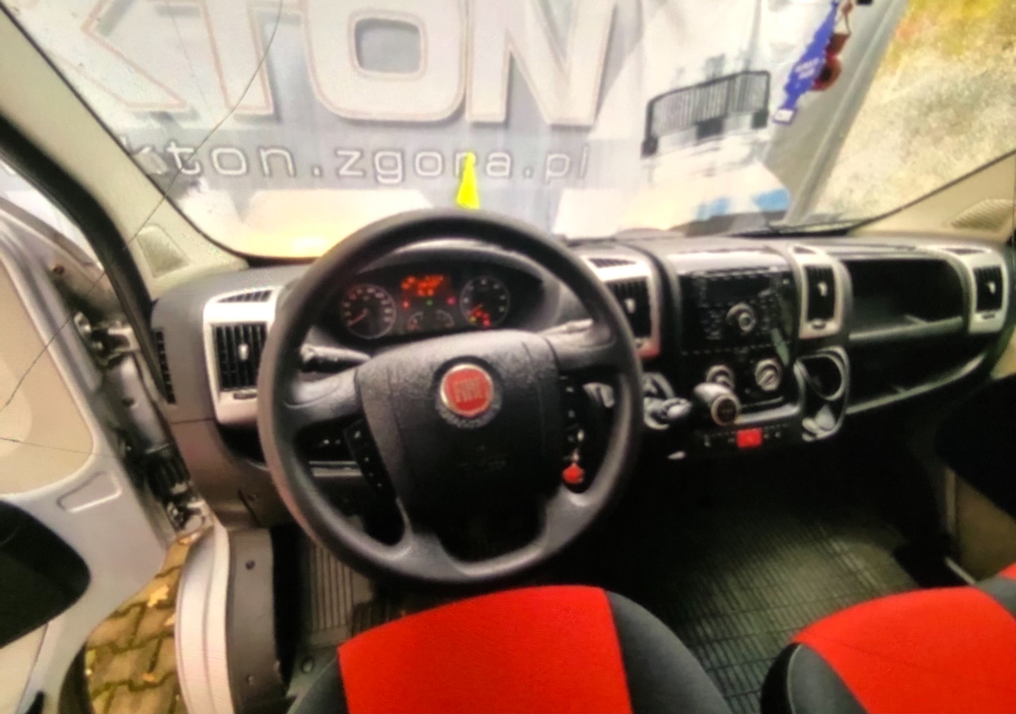Wnętrze samochodu dostawczego marki Fiat z widoczną deską rozdzielczą, kierownicą i czerwono-czarnymi pokrowcami na siedzeniach, z lekkim zniekształceniem obiektywu i odbiciem w szybie logotypu firmy.