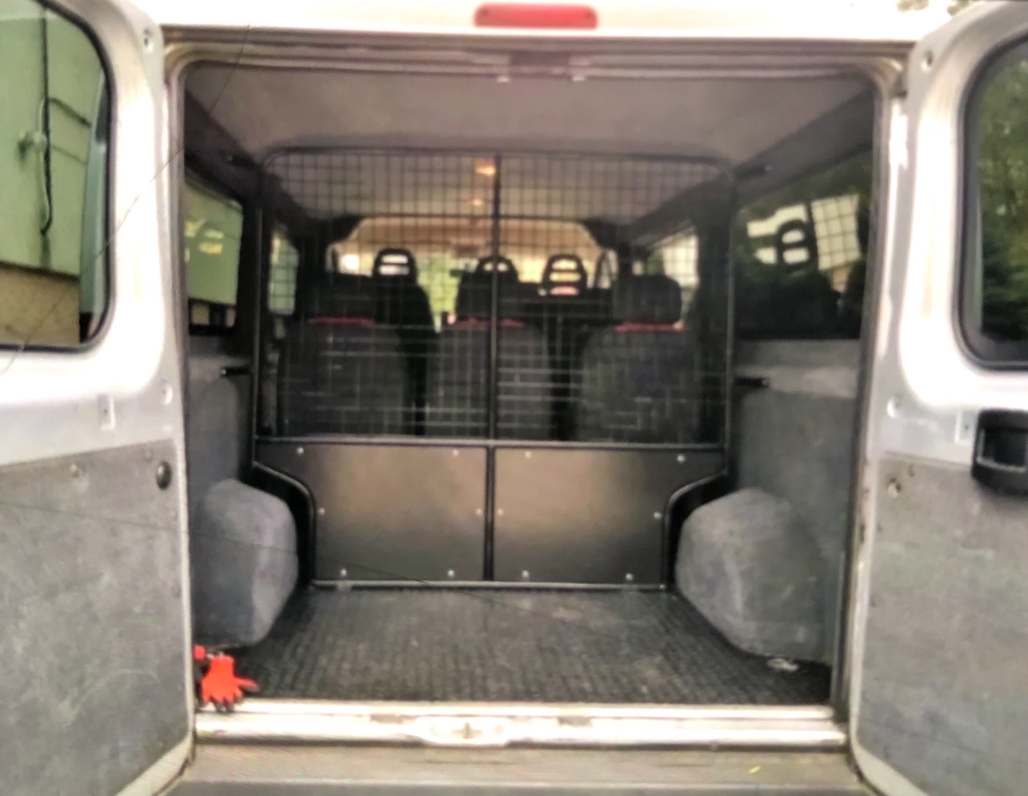 Wnętrze furgonetki z otwartymi tylnymi drzwiami, widoczna krata zabezpieczająca przestrzeń pasażerską, gumowa podłoga i para czerwonych rękawic roboczych w rogu.