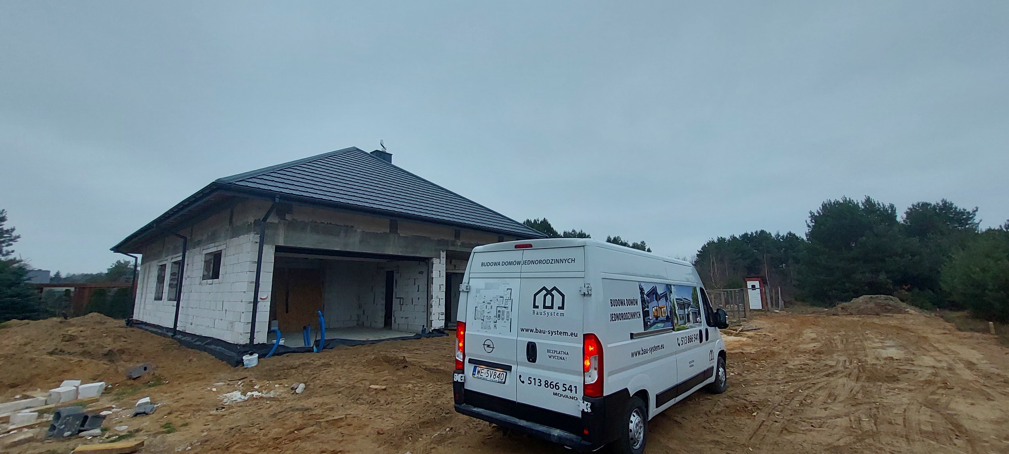 Niedokończony dom jednorodzinny z pustaków, z widoczną izolacją fundamentów i zaparkowanym obok samochodem dostawczym firmy budowlanej z reklamą na karoserii.