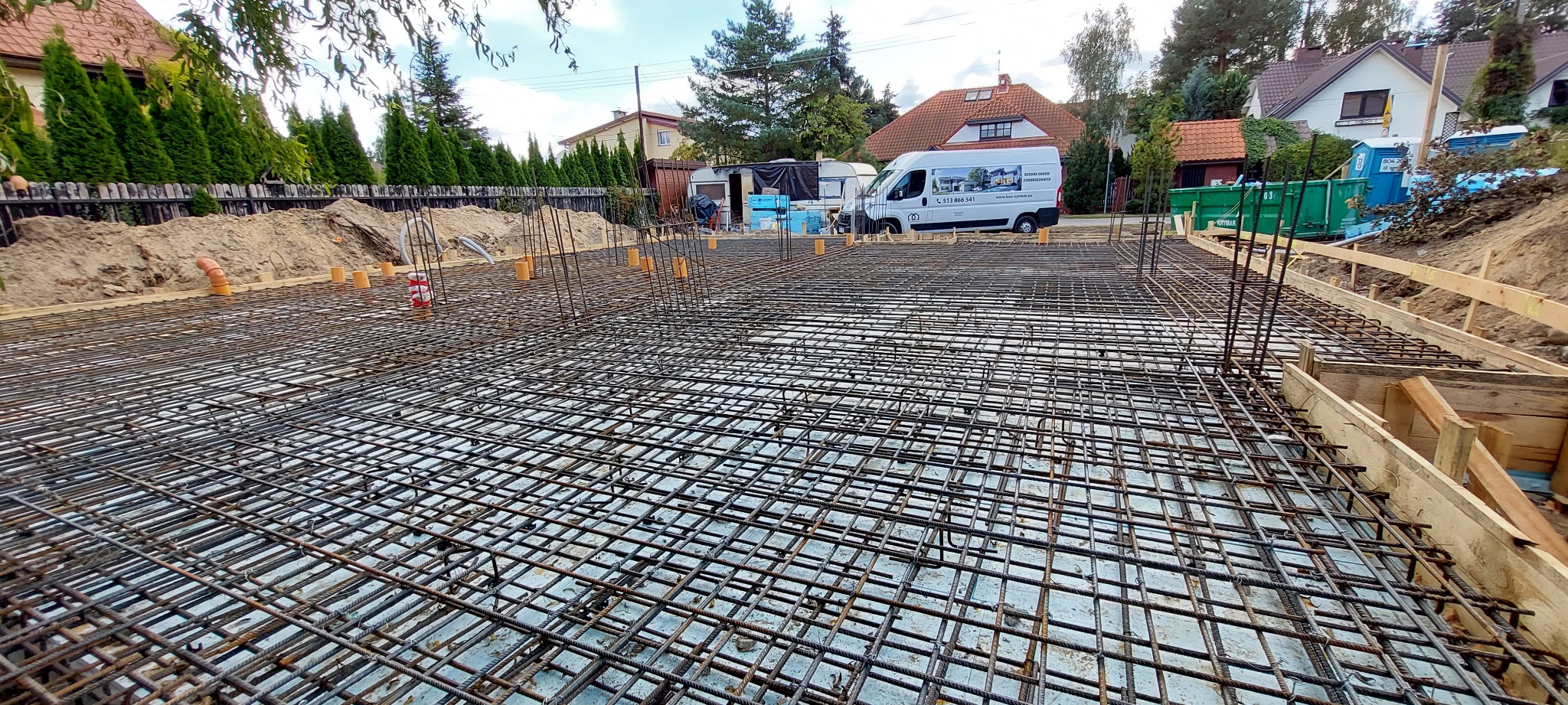 Zbrojenie płyty fundamentowej z wystającymi prętami pionowymi, szalunek drewniany po bokach, w tle domy jednorodzinne i firmowy bus.
