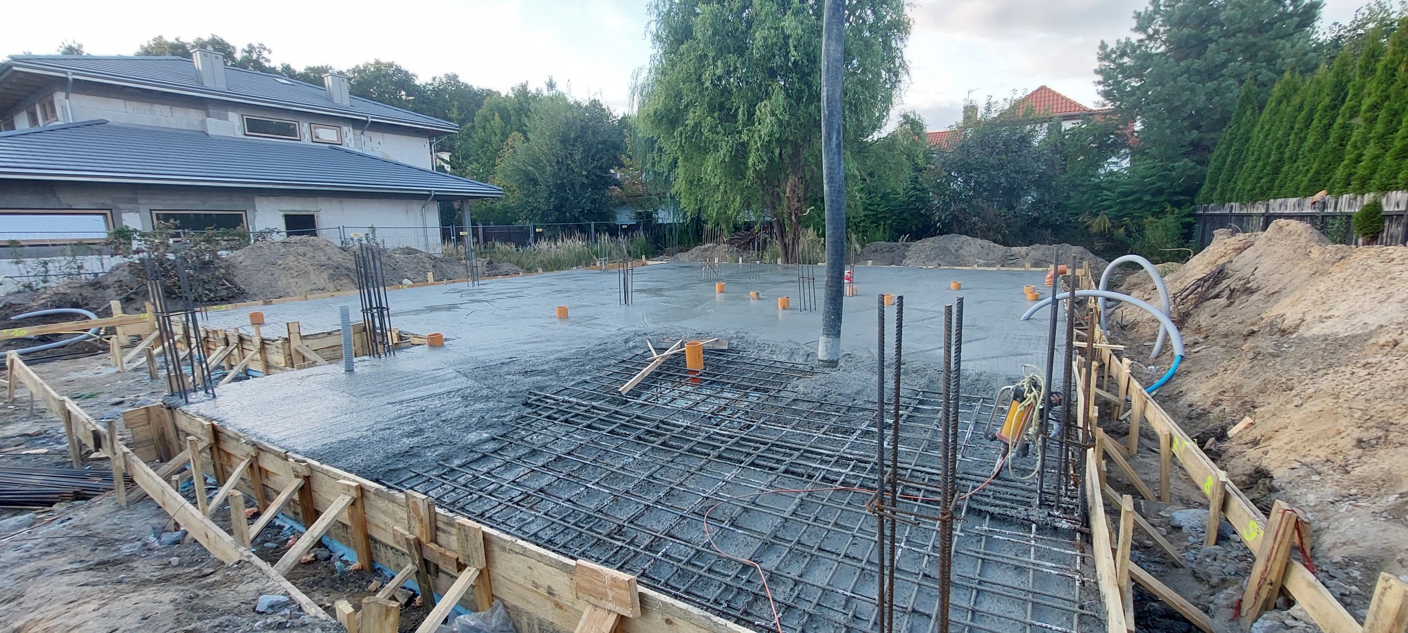 Wylewanie betonu na przygotowany szalunek zbrojeniowy pod budowę domu jednorodzinnego, widoczna pompa do betonu i wystające pręty zbrojeniowe.